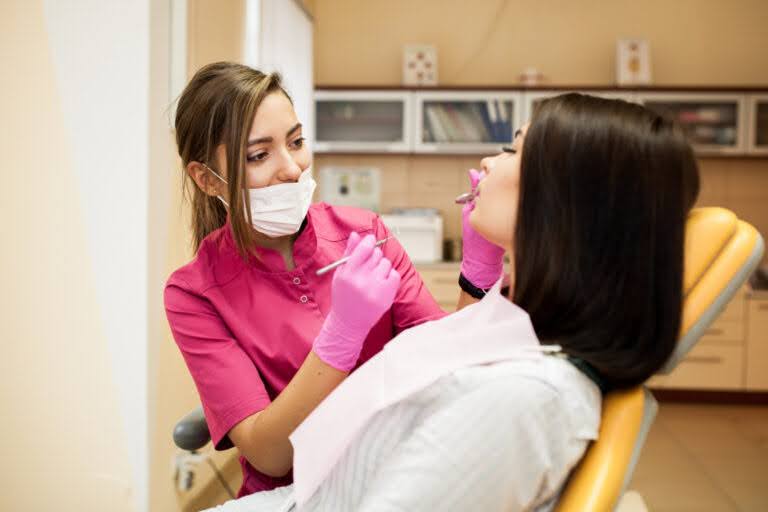 A female dentist in pink scrubs and a mask using a dental mirror to examine a patient's teeth in a clinic.